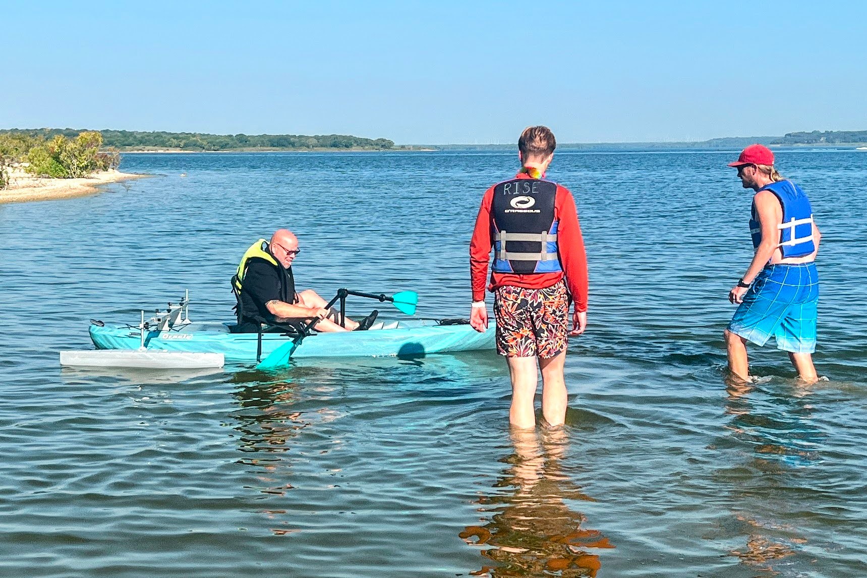 A kayaker uses a paddle support and outriggers while two people in life jackets stand nearby in Lake Grapevine.